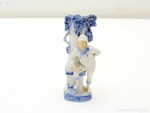 Charming porcelain boy figurine in sailor attire, playfully posed by a decorative grape column.