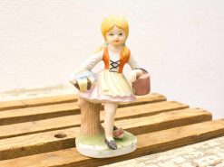 Charming porcelain figurine of a young girl with braids, basket, and pink bucket on a stump.