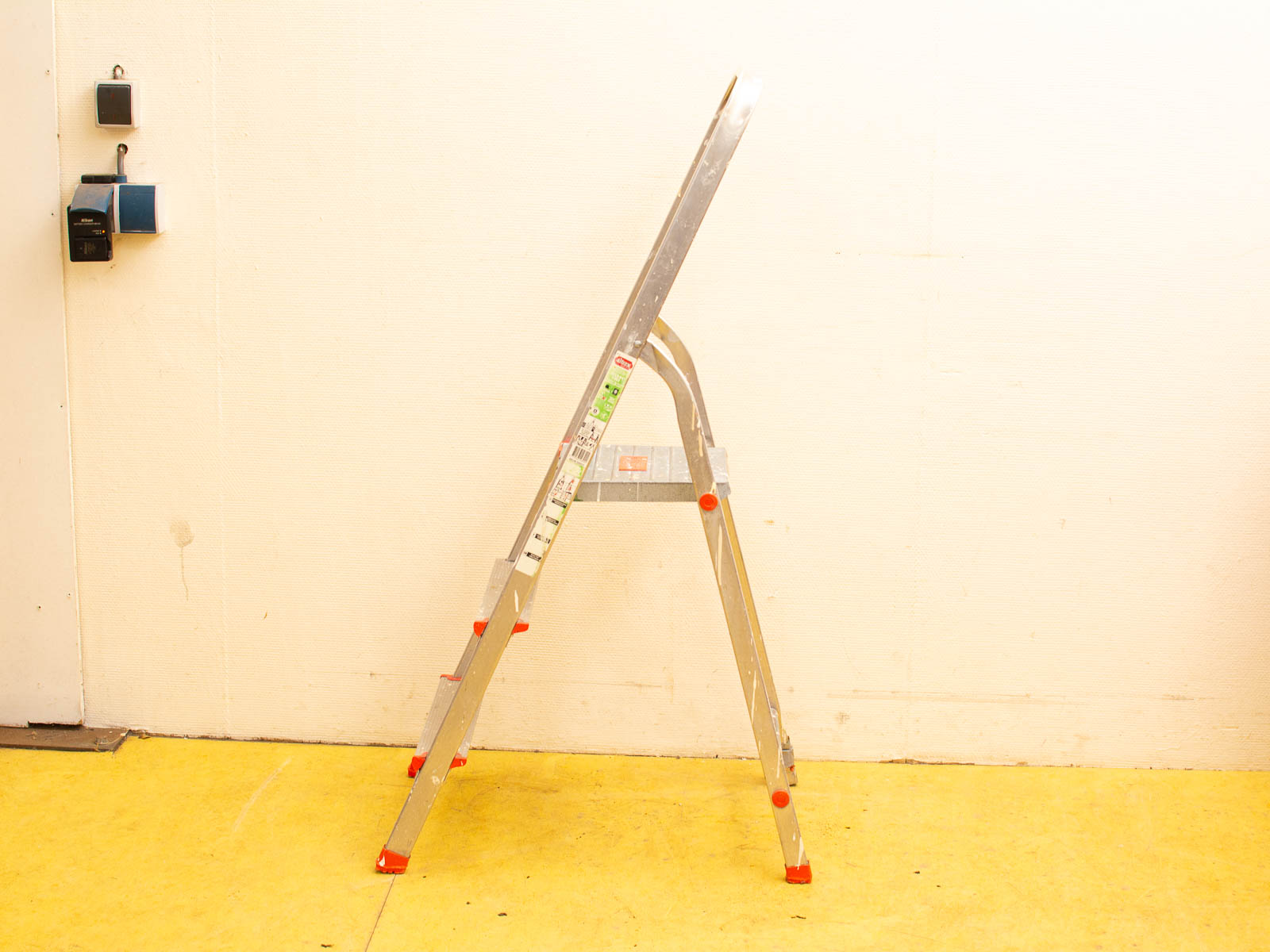 Aluminum folding ladder against textured wall on vibrant yellow floor in functional workspace.