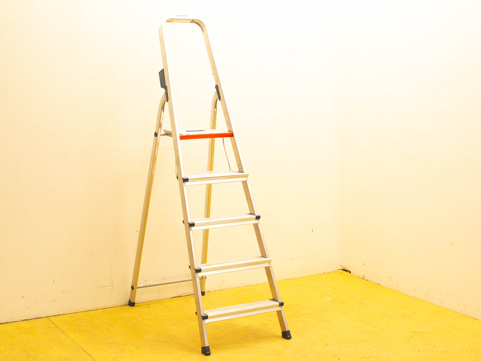 Modern aluminum step ladder with orange grip against a pale yellow wall, ideal for all tasks.