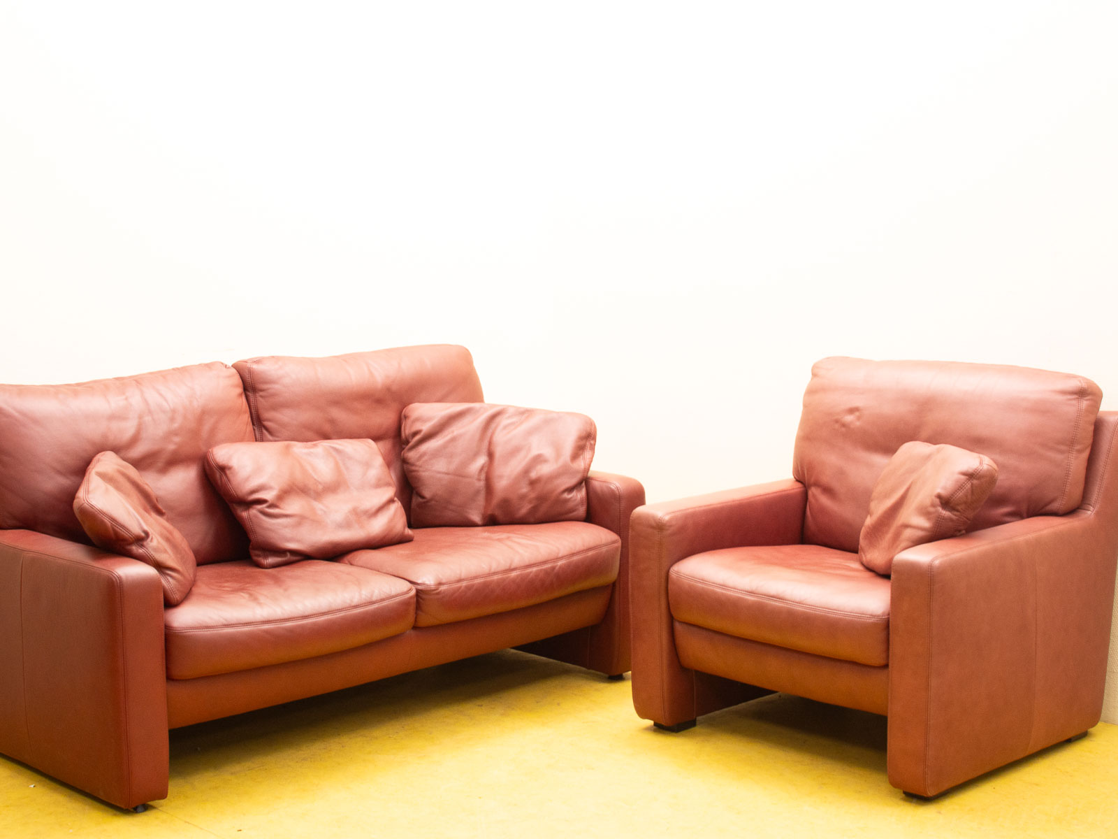 Cozy reddish-brown leather sofa and armchair on soft yellow carpet, perfect for modern living spaces.