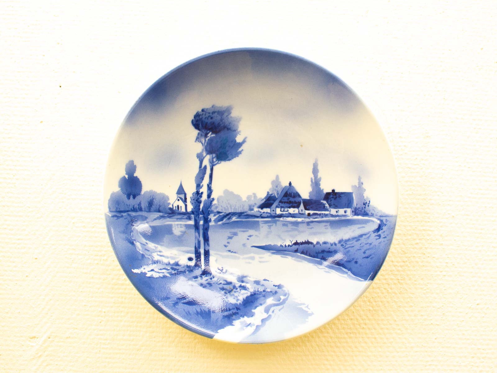 Serene blue decorative plate featuring a tranquil river, lush trees, and charming village homes.