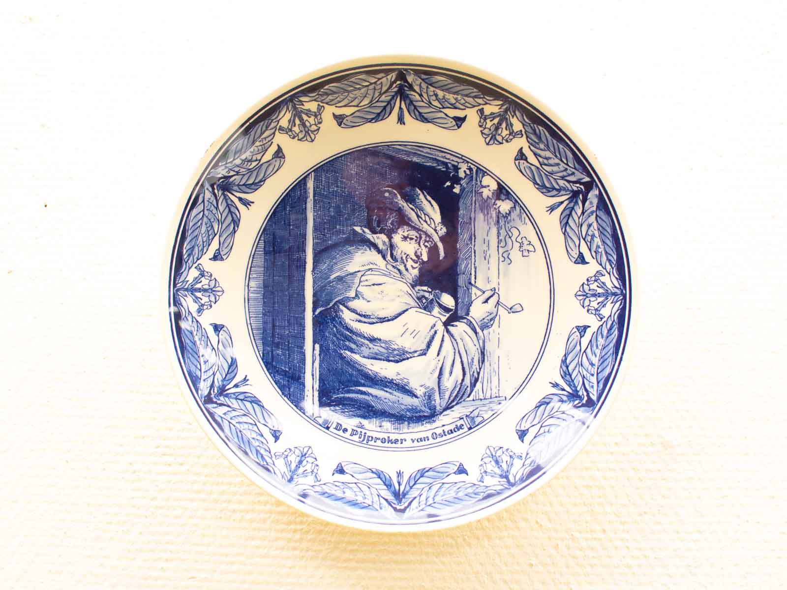 Vintage blue and white porcelain plate featuring The Pipe Smoker by Ostade, beautifully detailed.