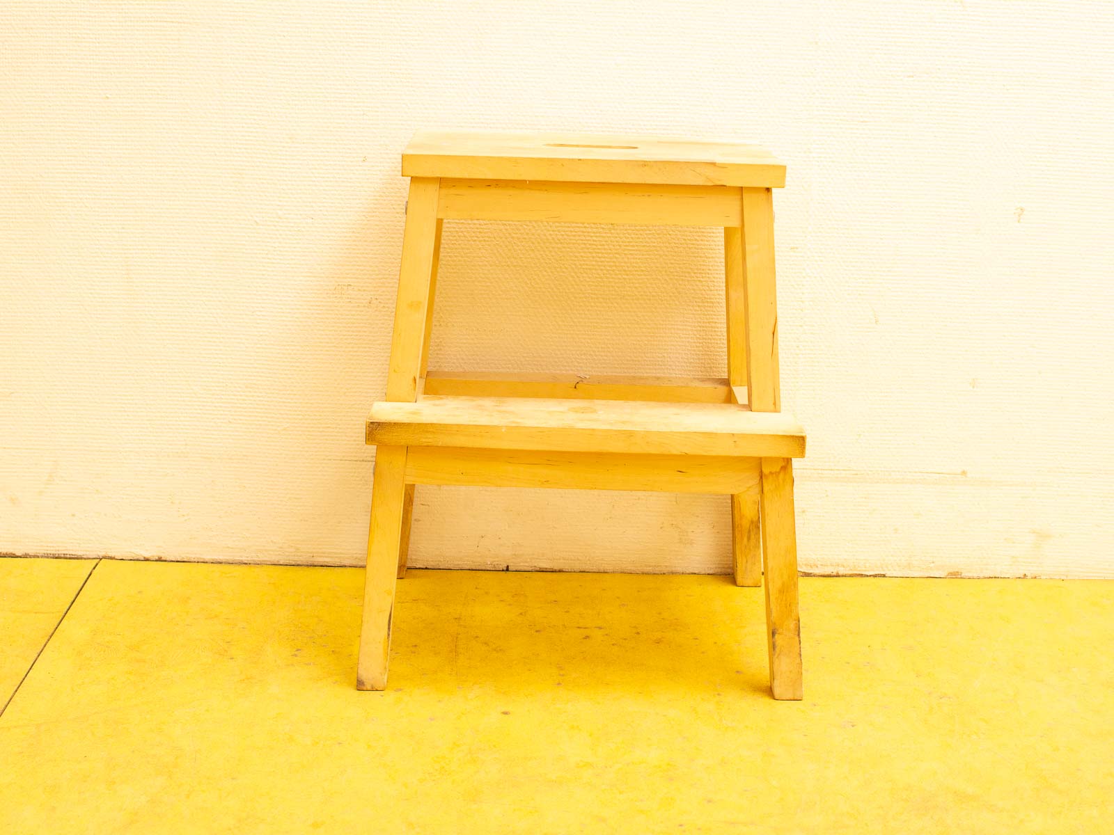 Light wooden step stool with perforated top against textured white wall and yellow floor.