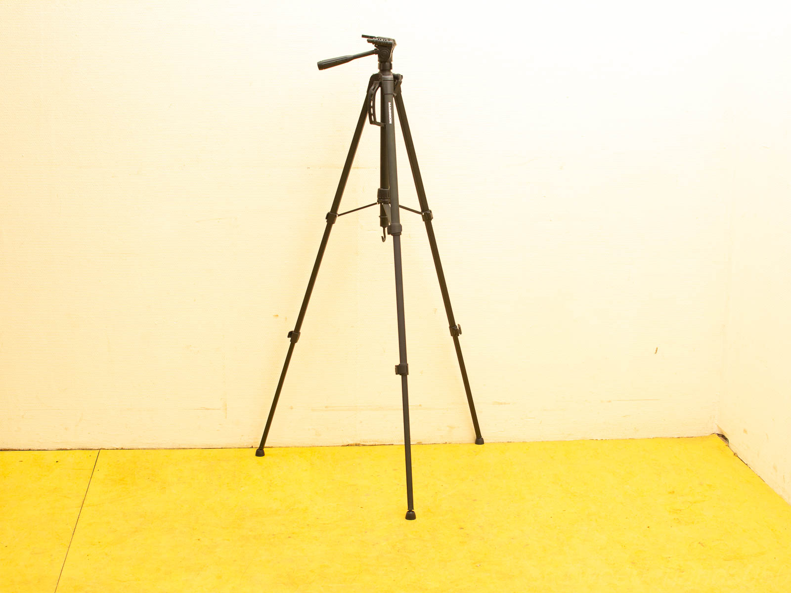 Black tripod on yellow floor, ideal for photography and videography with adjustable legs and ergonomic handle.