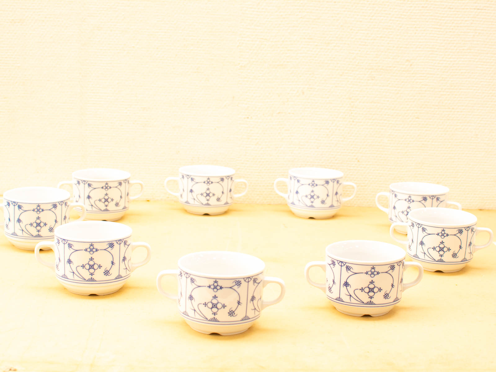 Ten elegant porcelain cups with blue floral patterns on a warm yellow surface.