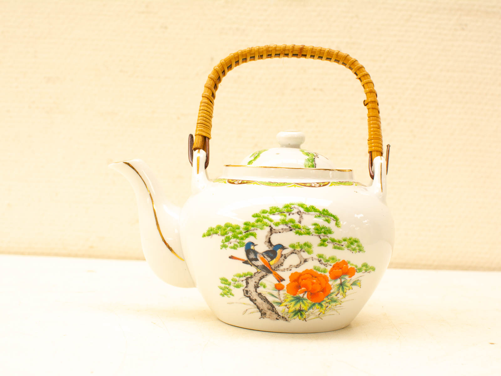 Vintage porcelain teapot with hand-painted flowers and a blue bird, featuring a woven rattan handle.