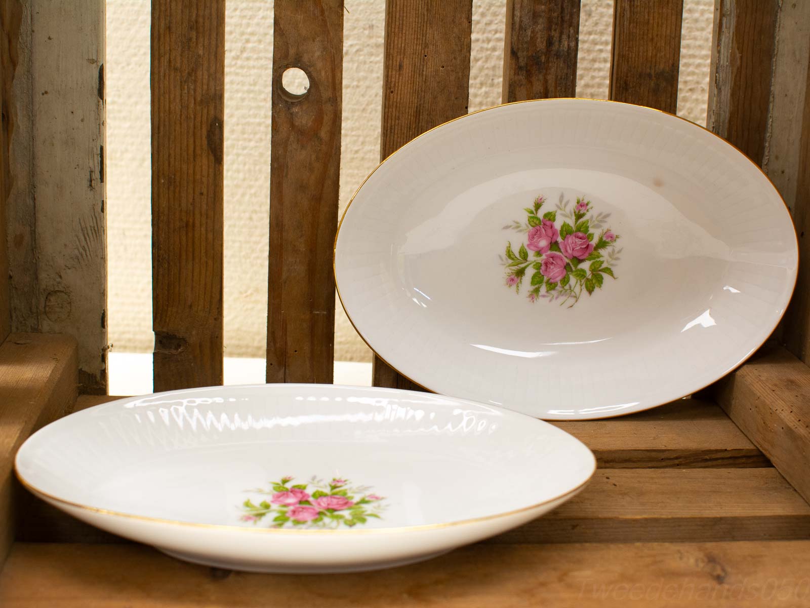 Elegant porcelain dishes with pink roses and gold trim on a rustic wooden shelf.