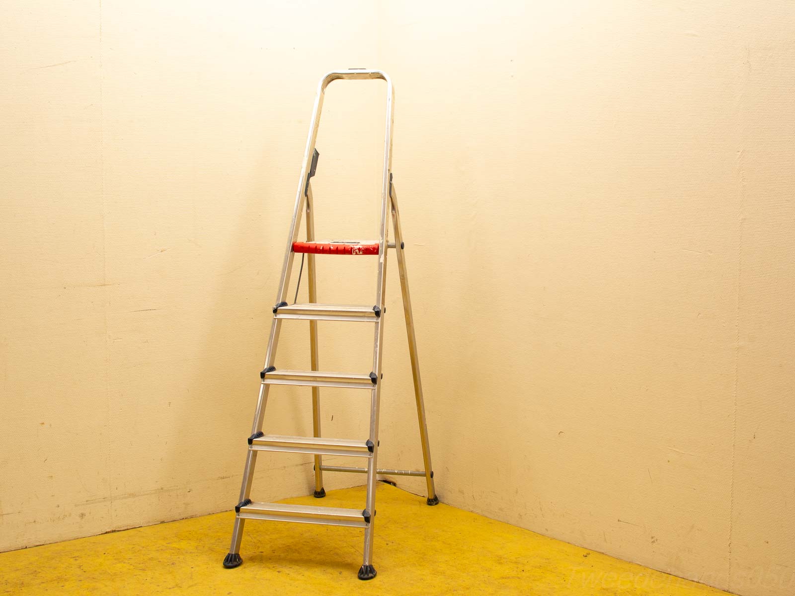 Sleek aluminum step ladder against a beige wall, featuring a vibrant red top step for safety.