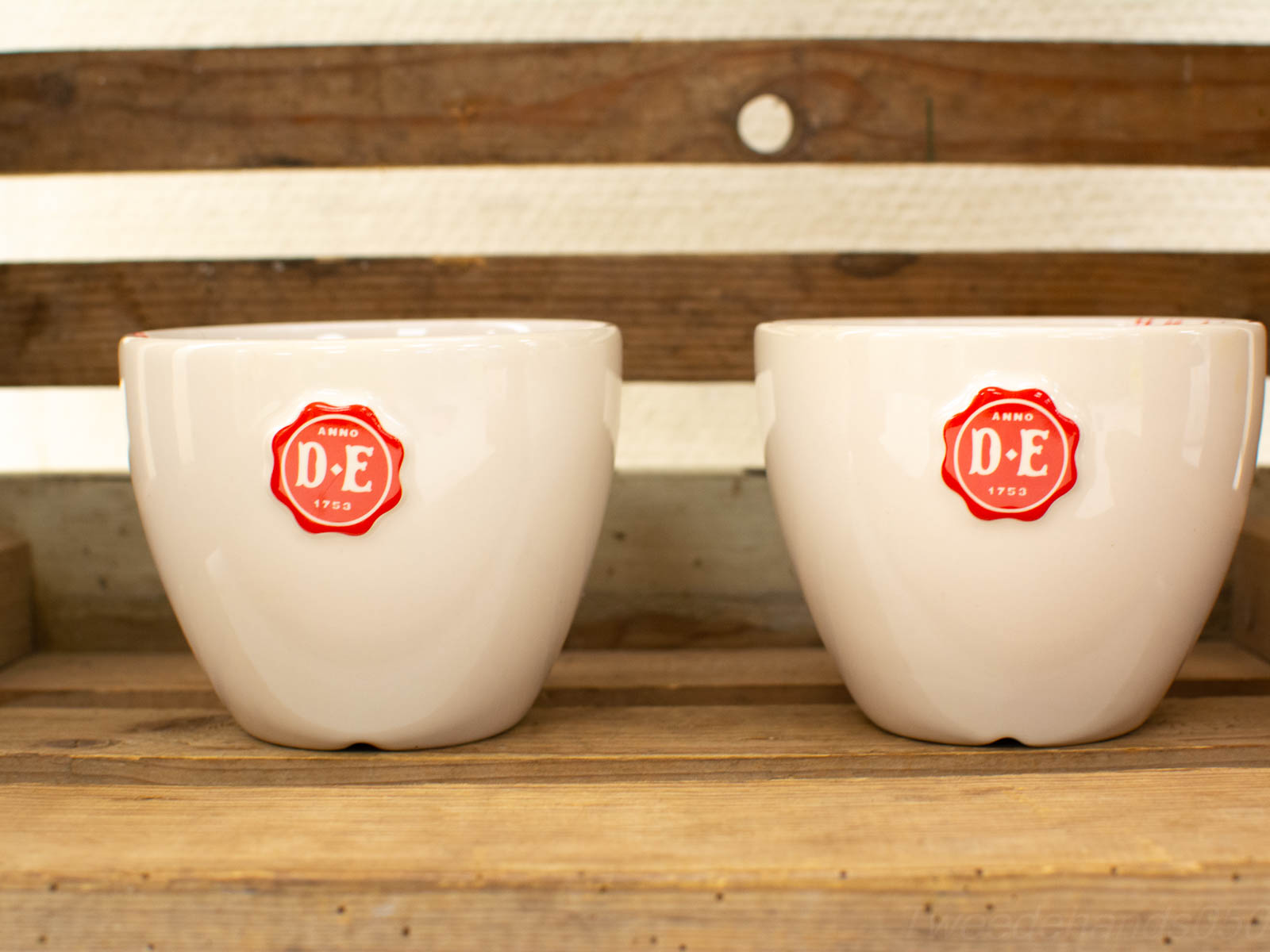 Elegant cream ceramic cups with red emblem on rustic wood, perfect for cozy gatherings.