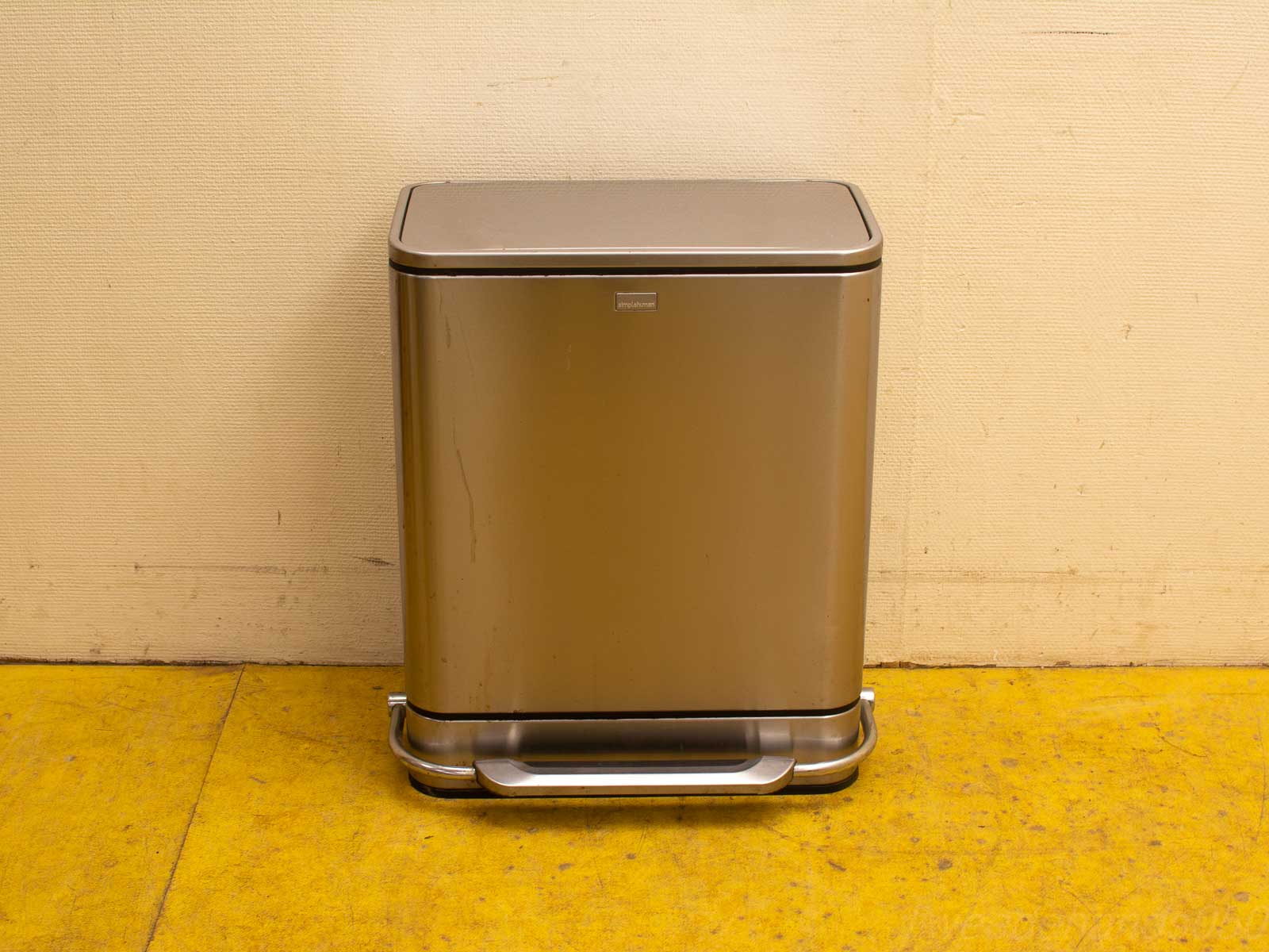 Modern stainless steel trash can with pedal against a textured beige wall and vibrant yellow floor.