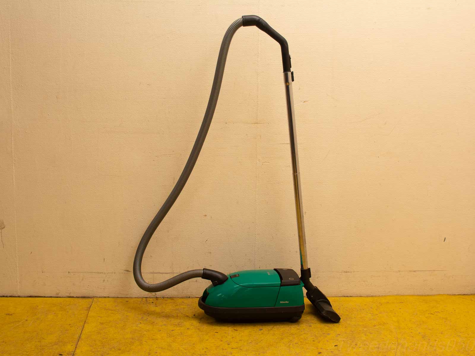 Sleek turquoise vacuum cleaner against a textured wall on vibrant yellow floor. Stylish and modern.