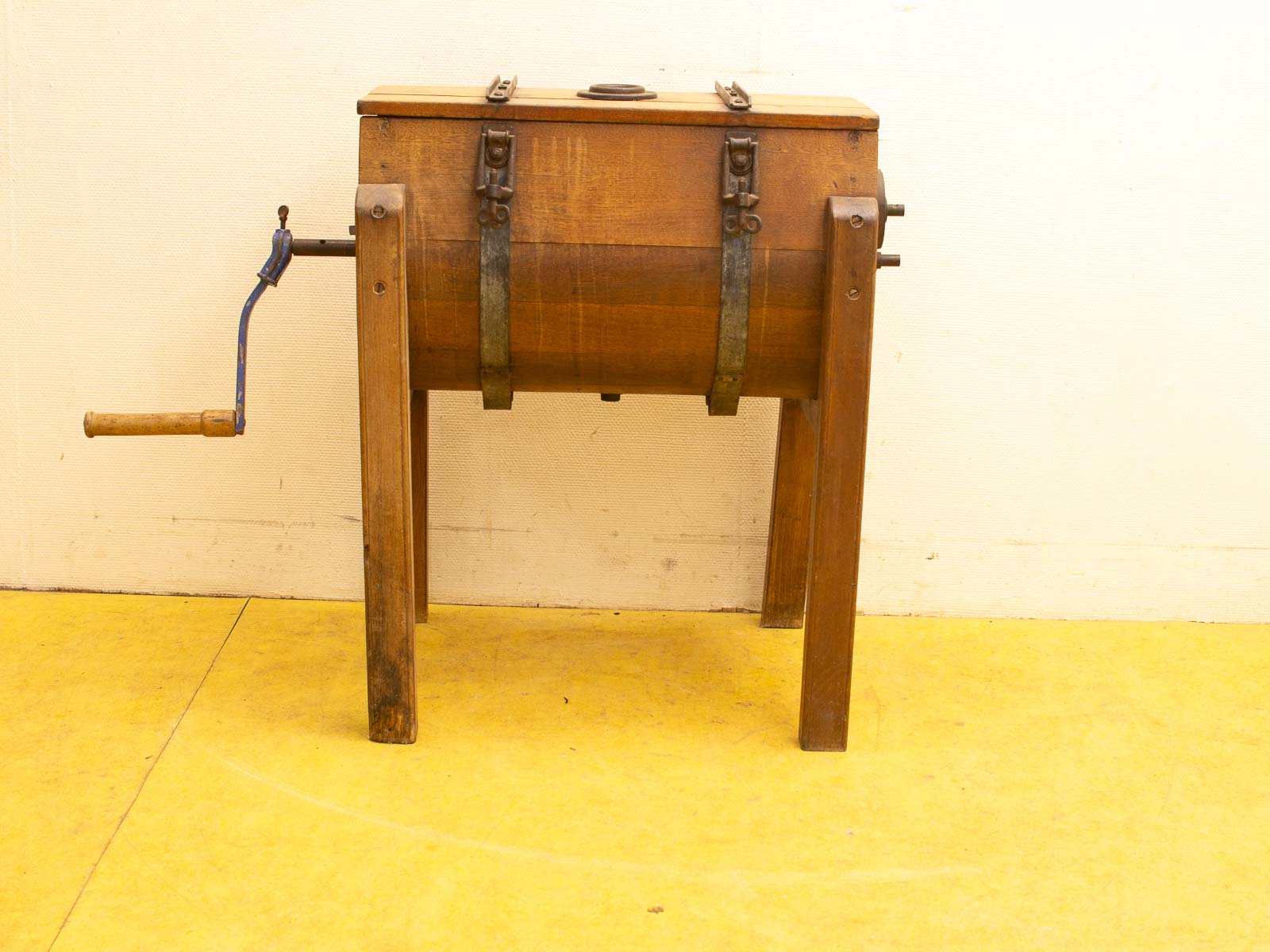 Vintage wooden butter churn with blue crank handle on a yellow floor, white wall background.