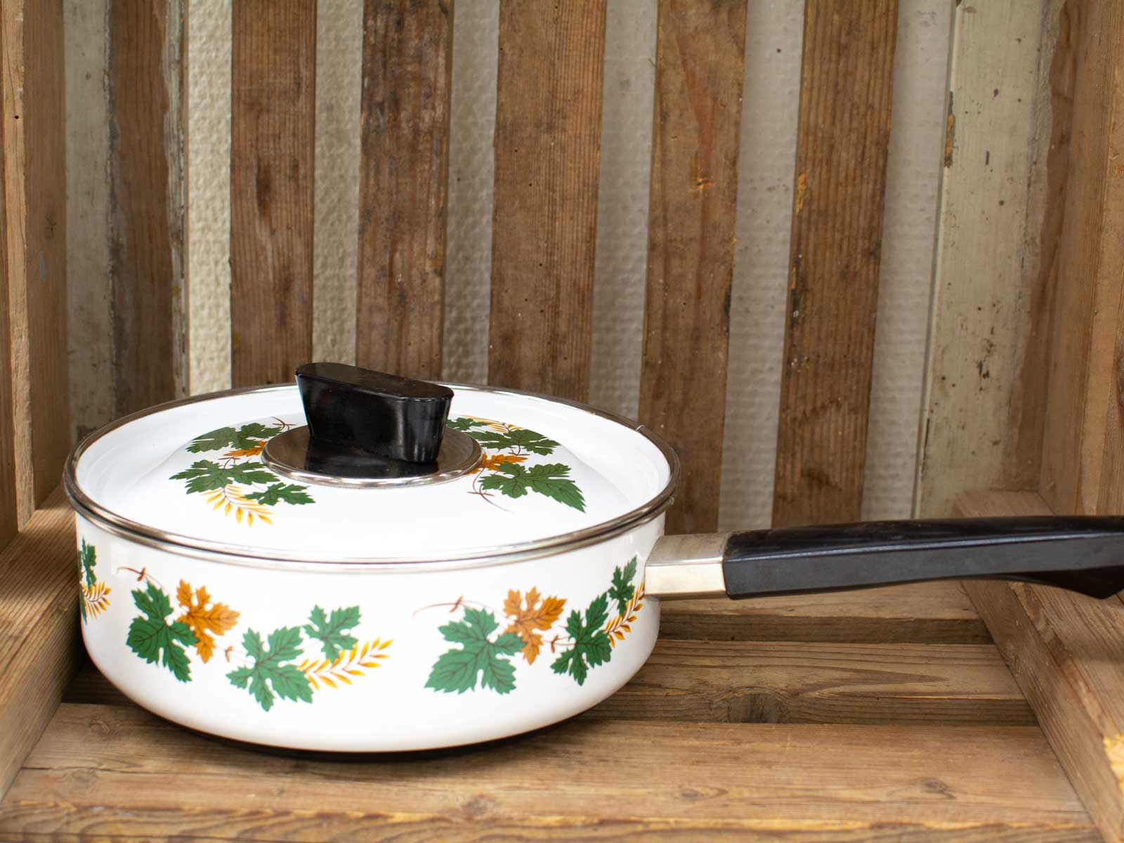 Vintage white saucepan with green and orange leaves on rustic wooden surface, perfect for home cooking.