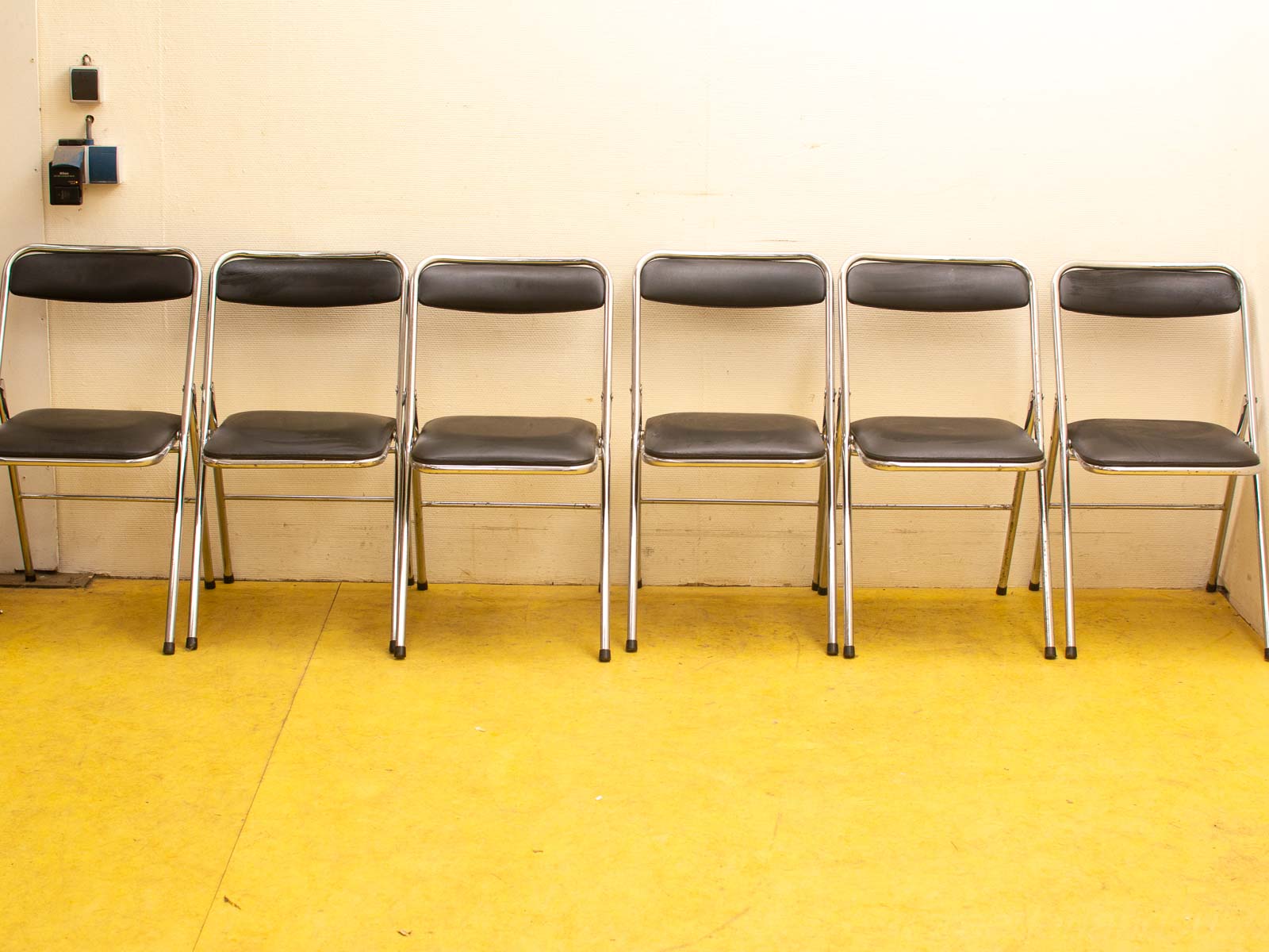 Six black minimalist folding chairs lined up on a vibrant yellow floor against a neutral wall.