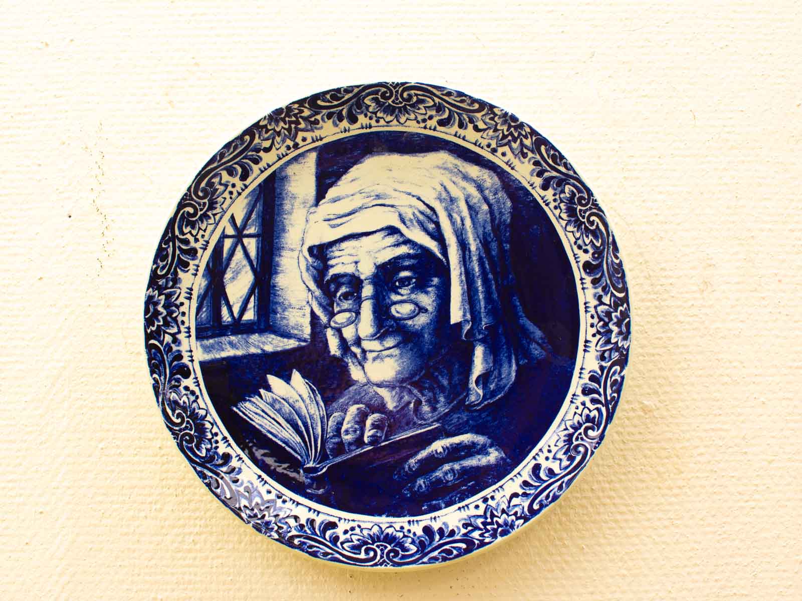 Vintage decorative plate featuring an elderly woman reading, beautifully framed in a blue floral border.