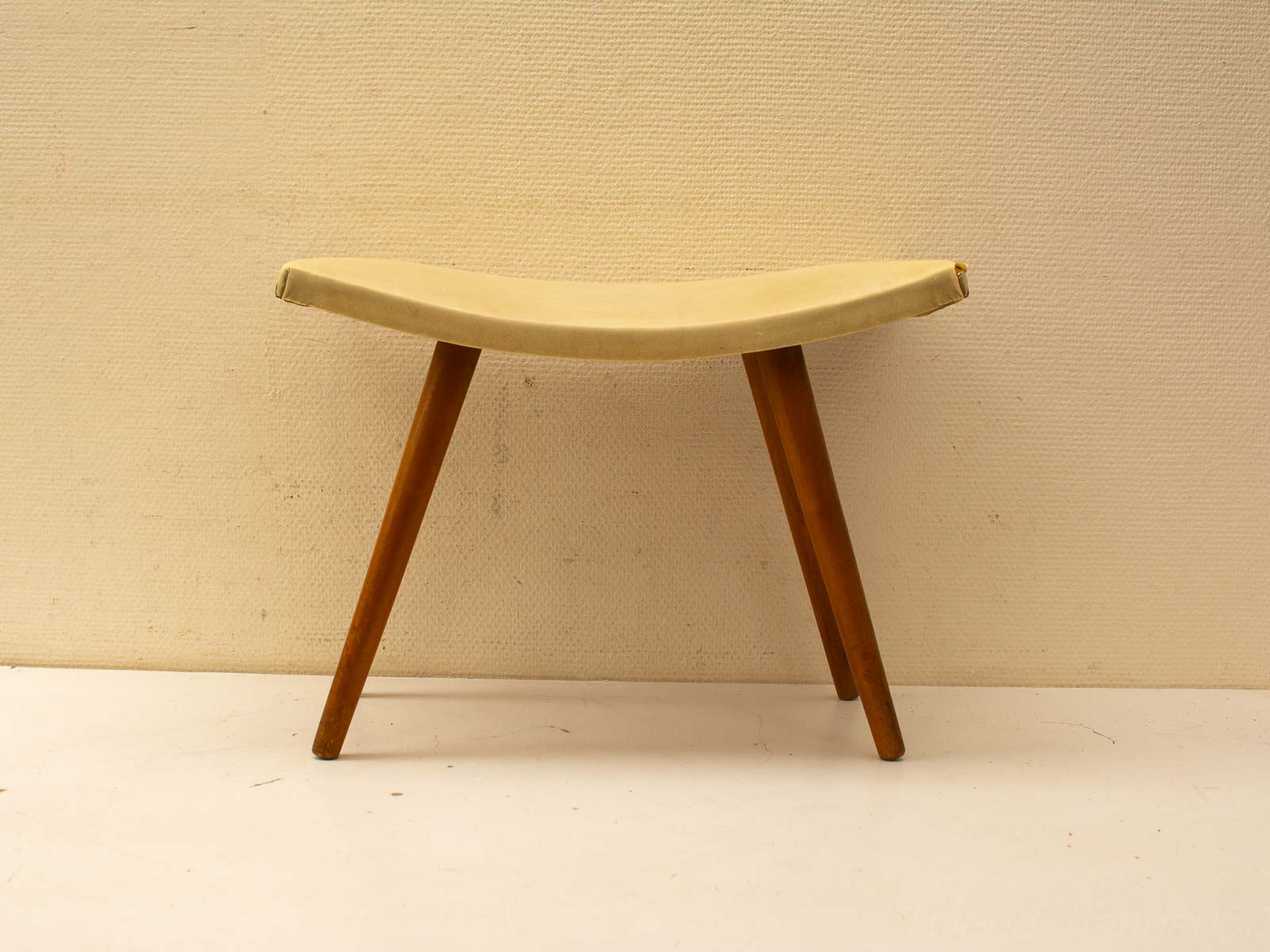 Elegant minimalist stool with curved beige seat and tapered wooden legs for modern interiors.