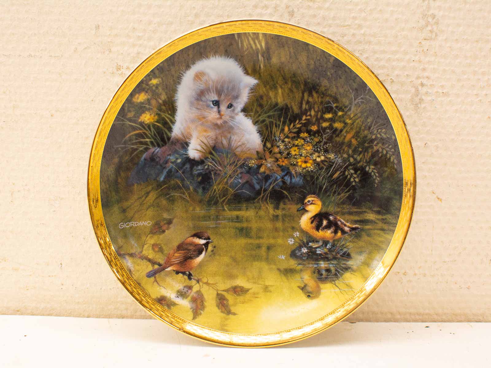 Fluffy white kitten and duckling by serene pond, vibrant nature scene on decorative plate.