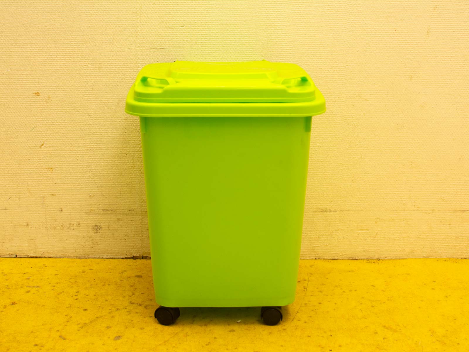 Heldergroene prullenbak met wielen op een gele vloer, ideaal voor recycling en afvalbeheer.