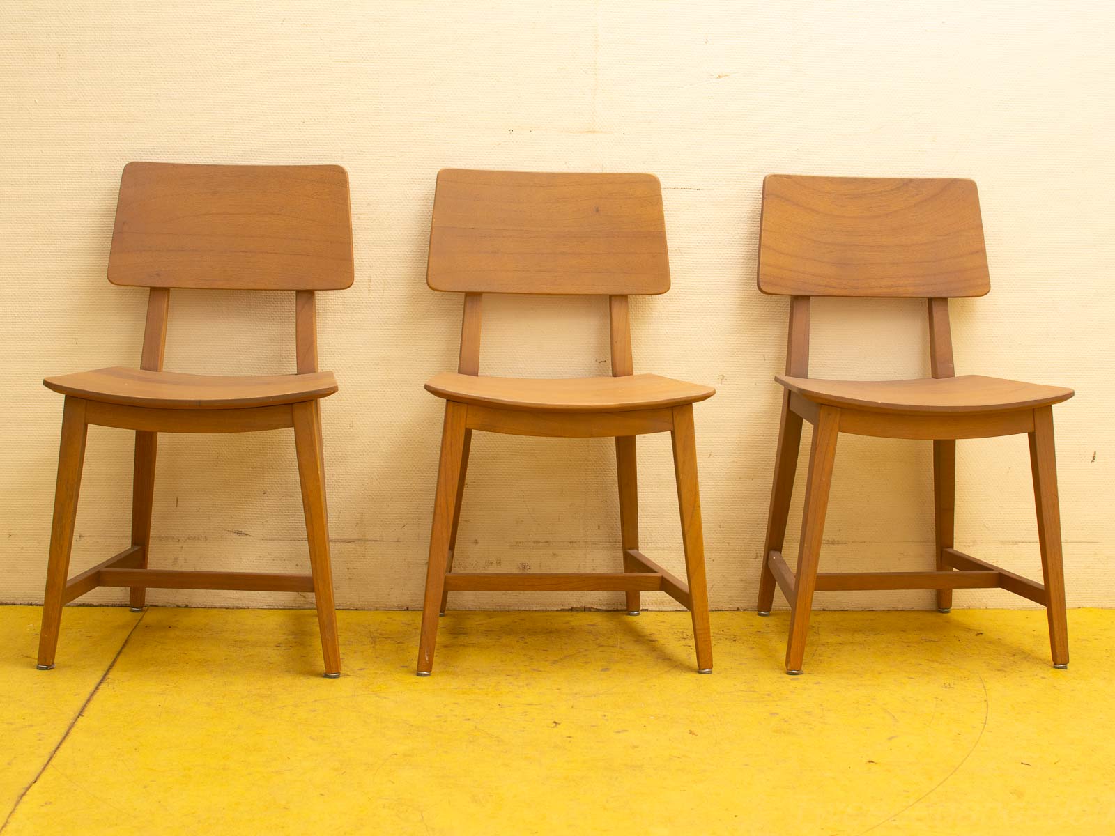 Three modern wooden chairs on a bright yellow floor against a light beige wall.