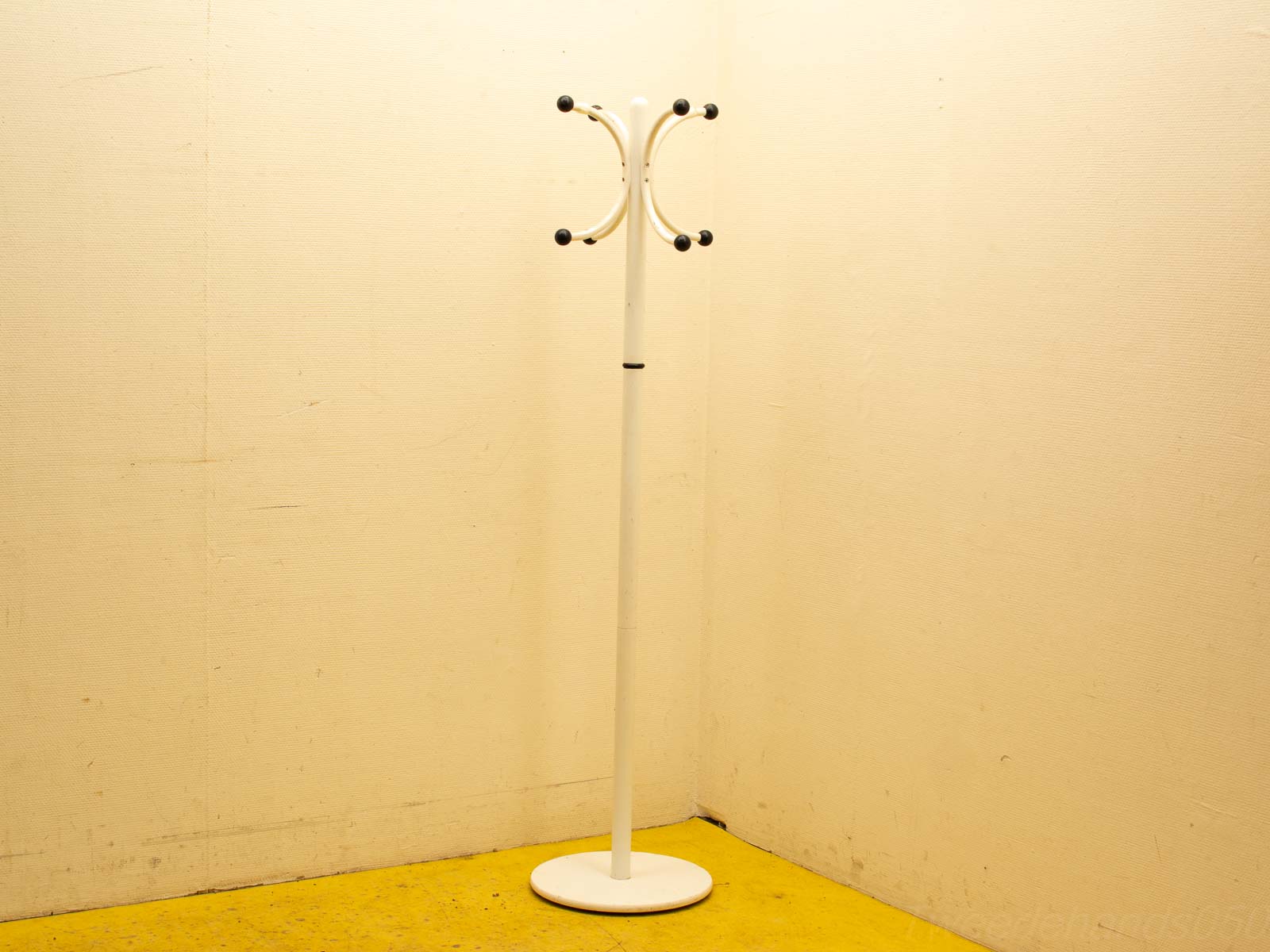 Sleek white coat rack with black-tipped hooks on a vibrant yellow floor against cream walls.