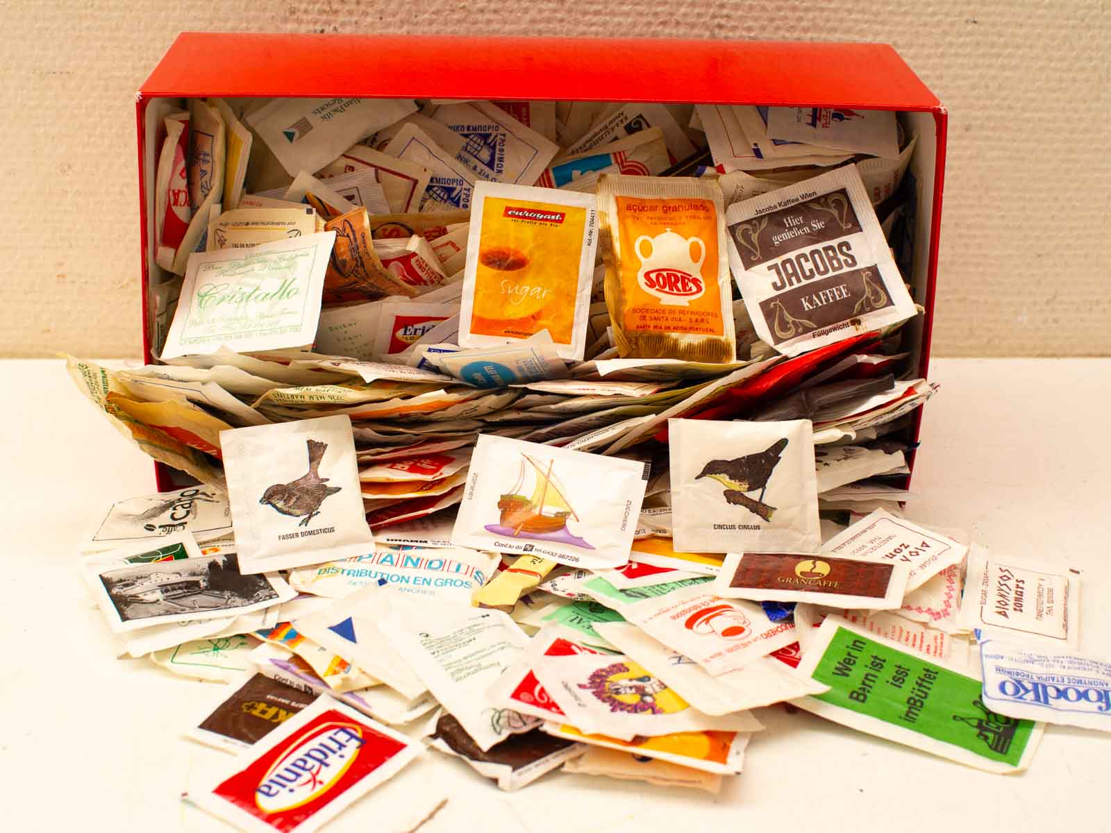 Vibrant red box overflowing with colorful vintage coffee and sugar packets, featuring unique designs.