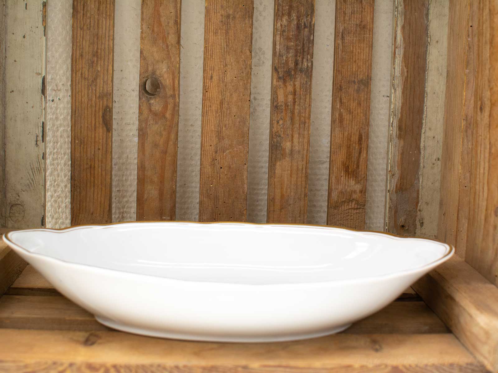 Elegant white porcelain dish with gold trim on rustic wooden shelf. Perfect decor for any setting.